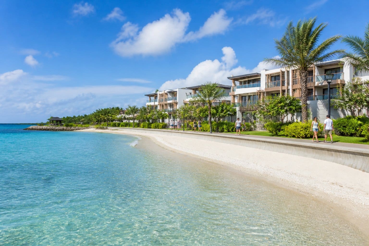 A beachfront neighborhood with modern houses, palm trees, clear blue ocean, and people walking along a promenade on a sunny day.