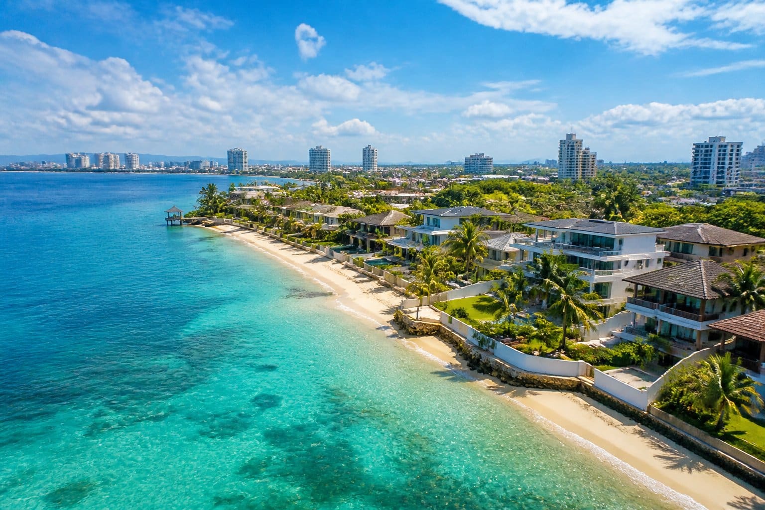 A coastal neighborhood with white sandy beaches, turquoise water, palm trees, and modern beachfront homes near a city skyline.