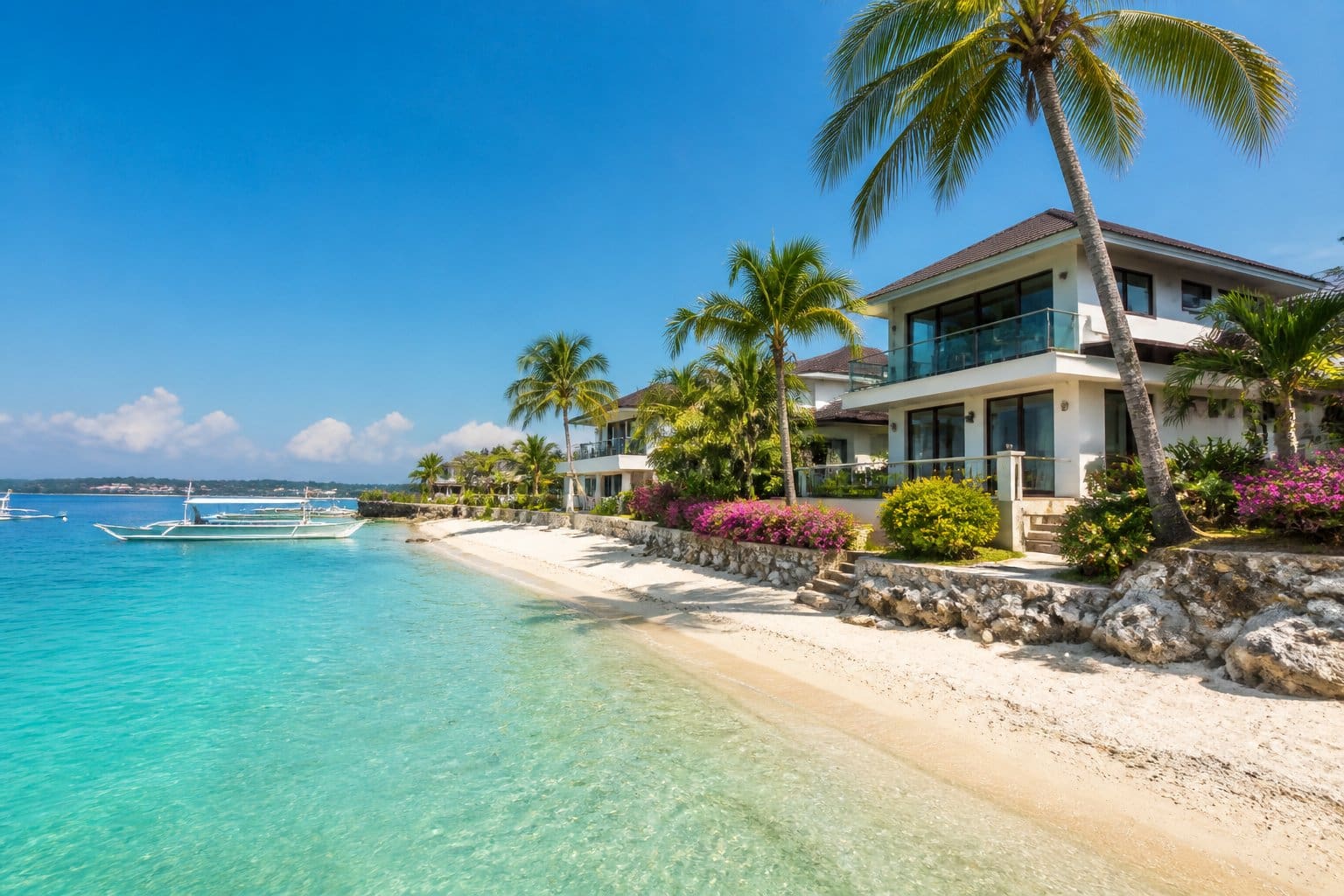 A beautiful beachfront neighborhood with modern houses, palm trees, and clear turquoise water under a bright blue sky.