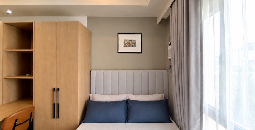 Bed with a grey upholstered headboard beside the oak wardrobe and built-in desk at Avida Towers Riala