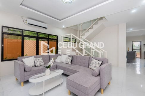Living room of the Binaliw house with a grey sectional sofa, aircon and glass-railed staircase