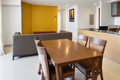 Wooden dining table with upholstered chairs set against a mustard accent wall beside the kitchen counter