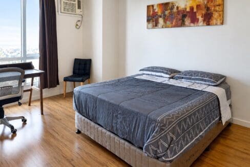 Main bedroom of this Lahug condo with wood-plank flooring, a desk by the window and a window-type air conditioner