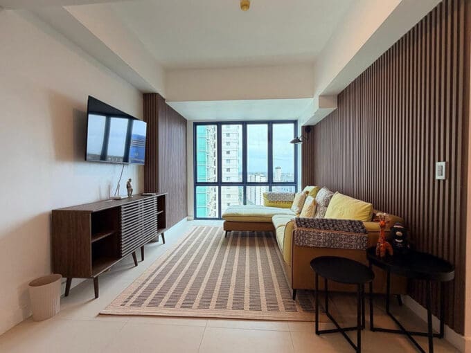 Living area with mustard L-shaped sectional, timber slat feature wall, and city views in the 2BR condo for rent at 38 Park Avenue