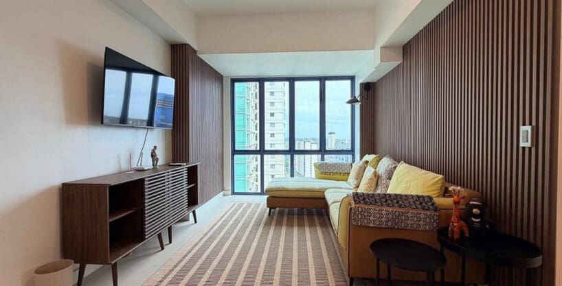 Living area with mustard L-shaped sectional, timber slat feature wall, and city views in the 2BR condo for rent at 38 Park Avenue