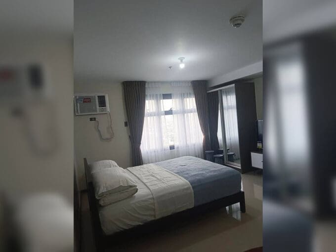 Sleeping area with window-type air conditioning and curtained window in the furnished studio at Galleria Residences