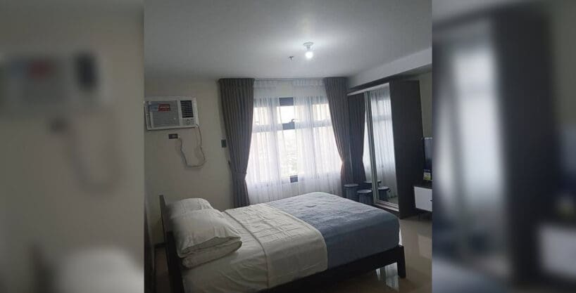 Sleeping area with window-type air conditioning and curtained window in the furnished studio at Galleria Residences