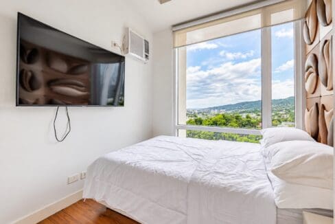 Bedroom with wall-mounted TV, window-type air conditioning and roller blind over the picture window