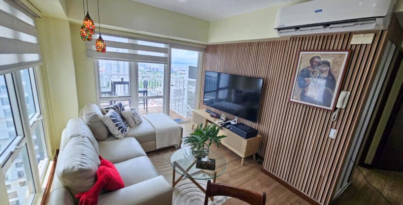 Living area with cream sectional sofa, slatted timber TV wall and balcony door in a 2BR condo for sale at Solinea Tower 2, Cebu Business Park