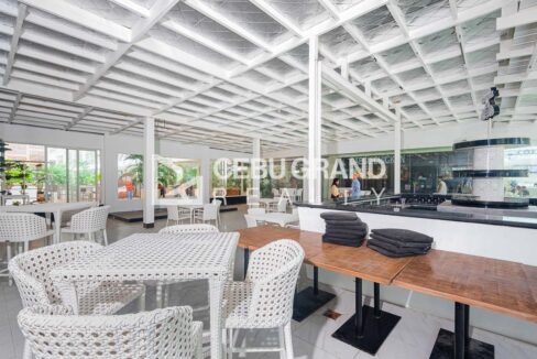 Commercial space with white rattan dining sets, timber communal tables and a black-top bar counter beside glass-fronted food stalls