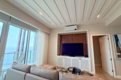 TV set in a slatted wood niche above louvered cabinets, with the sea beyond the glass